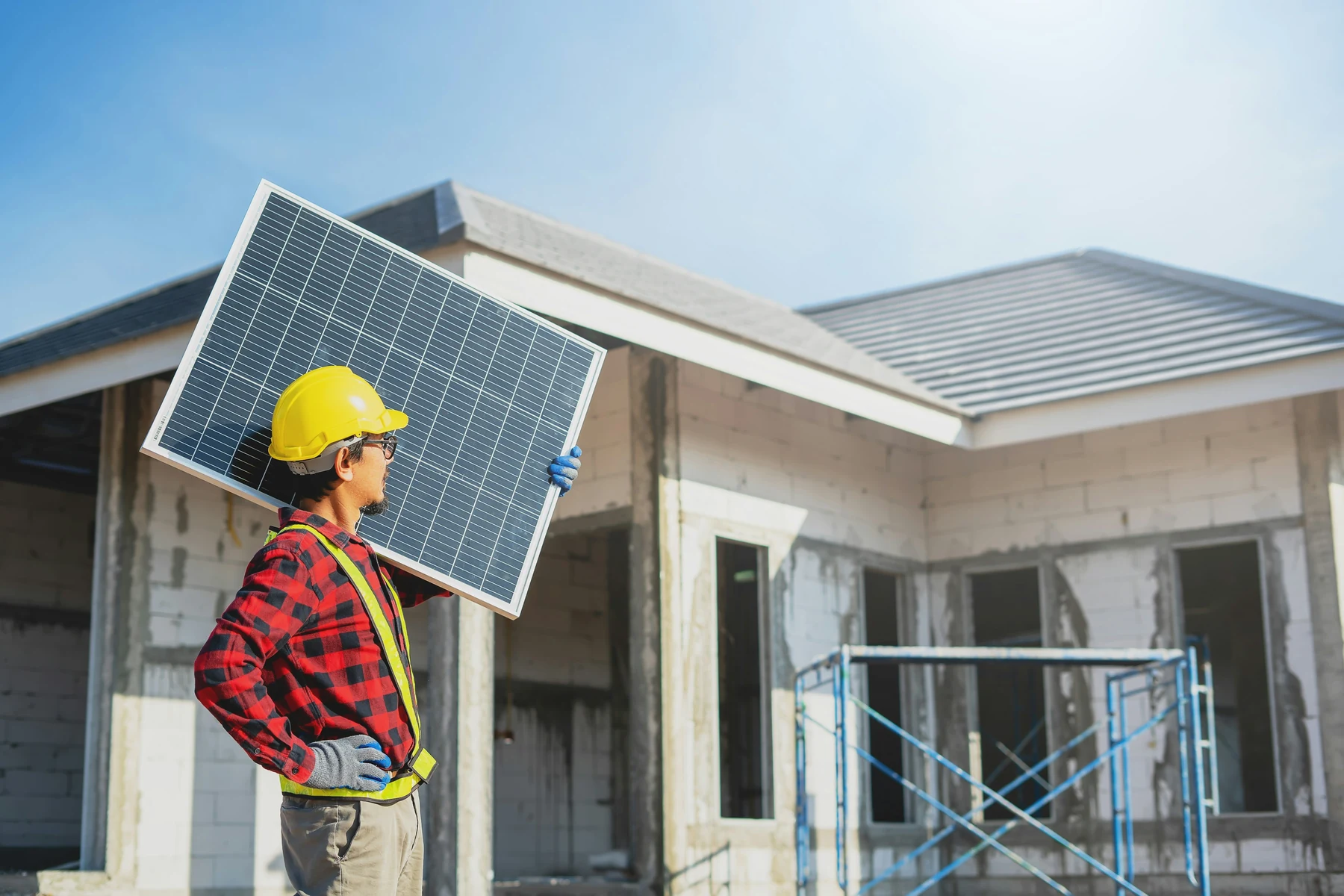 Installateur casqué transportant un module photovoltaïque devant une maison en chantier